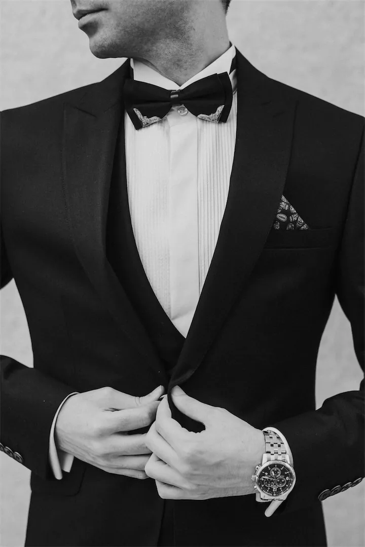 Man buttoning a black tuxedo with bow tie and pocket square
